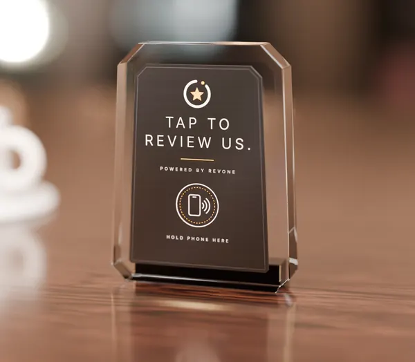 A Revone crystal acrylic standee on a café counter reading Tap to review us, with a phone tap ring.
